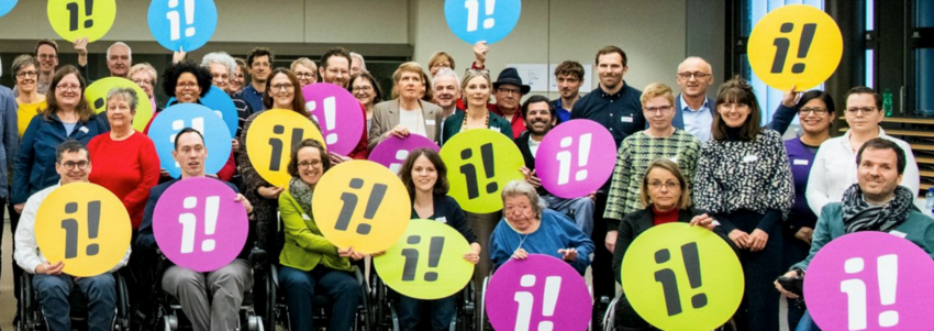 Eine Gruppe von Menschen mit Behinderung mit farbigen Plakaten mit einem i und einem Ausrufezeichen